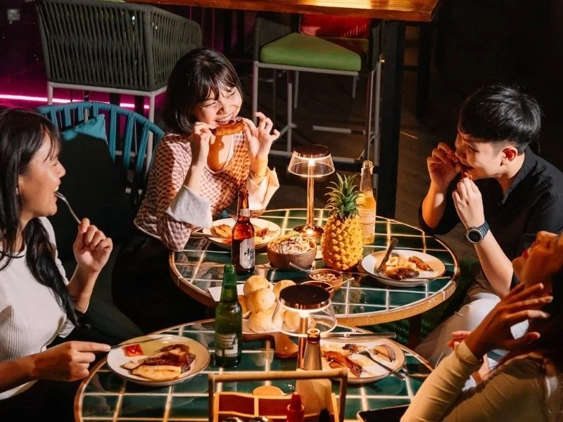 Friends sharing Thai-inspired dishes, a pineapple and drinks at a tiled table on Mimi’s rooftop at night