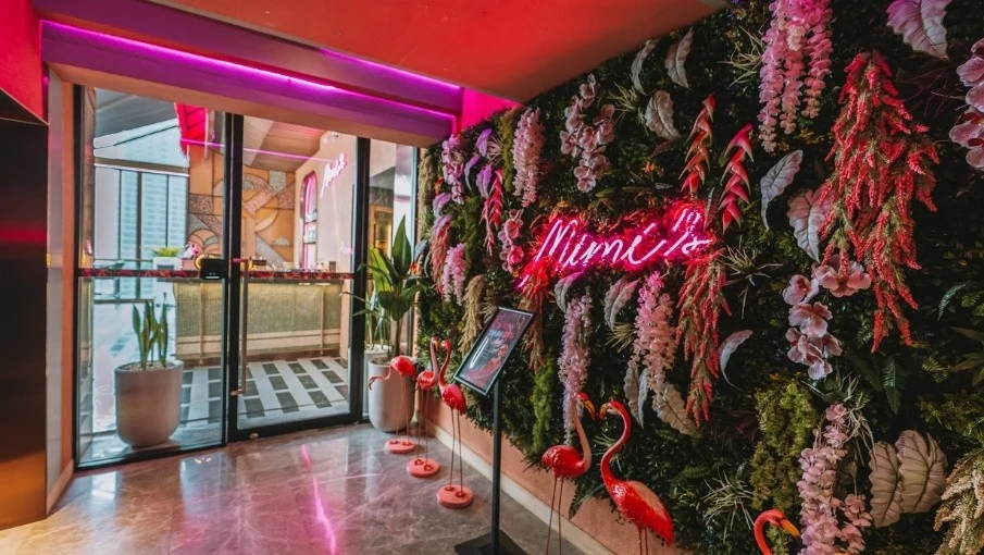 Entrance to Mimi’s with a pink neon Mimi’s sign on a tropical flower wall and flamingo figures