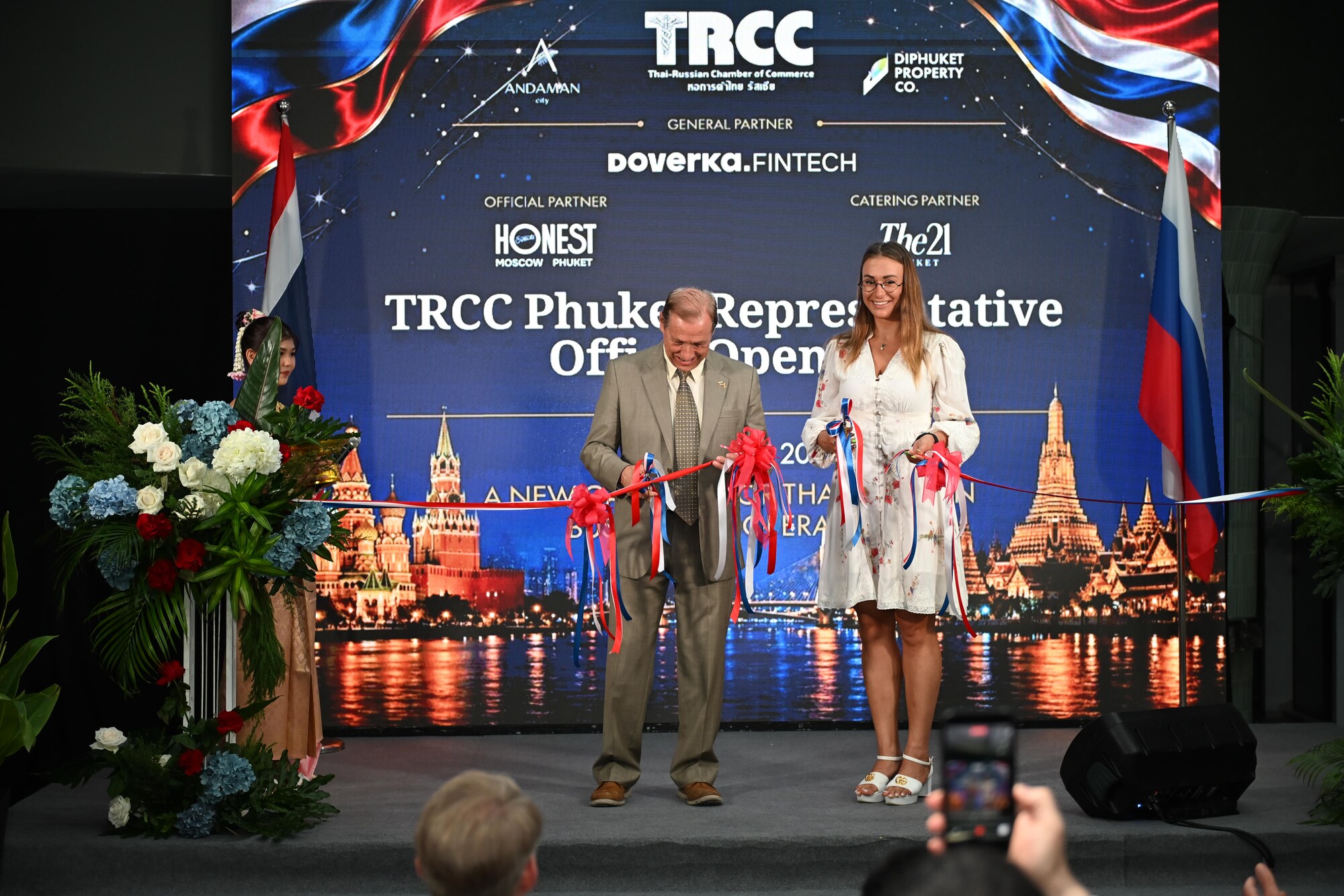 A man in a suit and a woman in a white dress cutting the red opening ribbon.